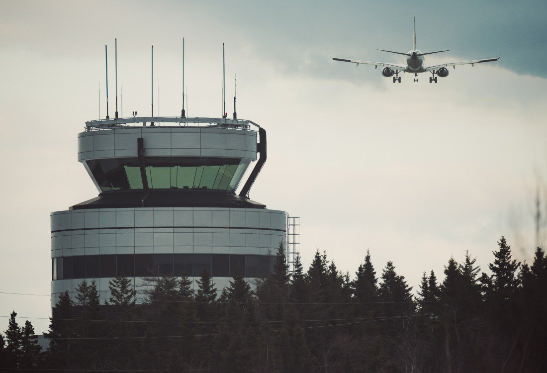 An air traffic control tower surrounded by trees with an airplane approaching for landing in the background, symbolizing the importance of ATC systems and safety redundancy in global flight security. An air traffic control tower surrounded by trees with an airplane approaching for landing in the background, symbolizing the importance of ATC systems and safety redundancy in global flight security.